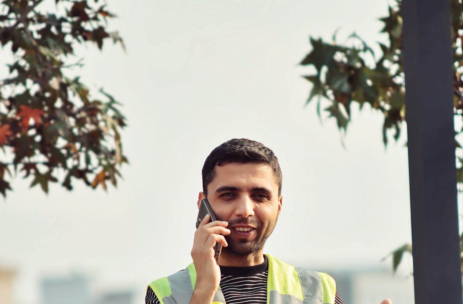 Handwerker in Warnweste telefoniert im Freien mit dem Smartphone am Ohr und laechelt
